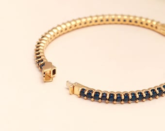 May include: Gold-coloured bracelet with a row of small, dark blue gemstones. The bracelet is open, showing the clasp mechanism. The gemstones are evenly spaced along the band, creating a continuous line of colour.