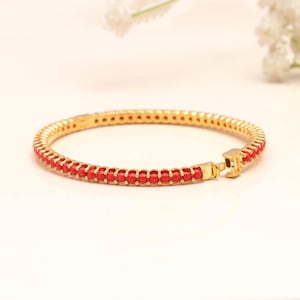 Puede incluir: Una pulsera rígida de tono dorado que presenta una hilera continua de pequeñas cuentas esféricas de color rojo vibrante engastadas en un marco metálico. La pulsera incluye un mecanismo de cierre de bisagra seguro visible en el lado derecho.