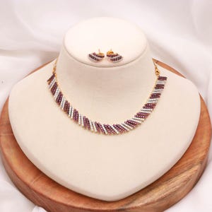 Puede incluir: Un conjunto de collar y pendientes a juego en tono dorado con un patrón diagonal repetitivo de piedras blancas transparentes y color magenta intenso. Las piezas de joyería presentan un diseño lineal estructurado con filas alternas.