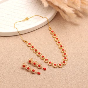 Puede incluir: Un conjunto de collar y pendientes dorados. Las joyas presentan un diseño floral con piedras centrales rojas y detalles blancos. Los pendientes tienen una piedra roja en forma de lágrima en la parte inferior.