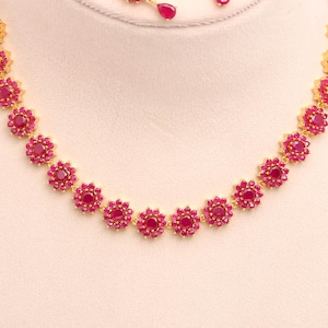 May include: A gold necklace with a floral pattern, set with bright red gemstones. The necklace is made up of flower-shaped settings, each with a central red stone and smaller red stones around it. Matching earrings are also visible.