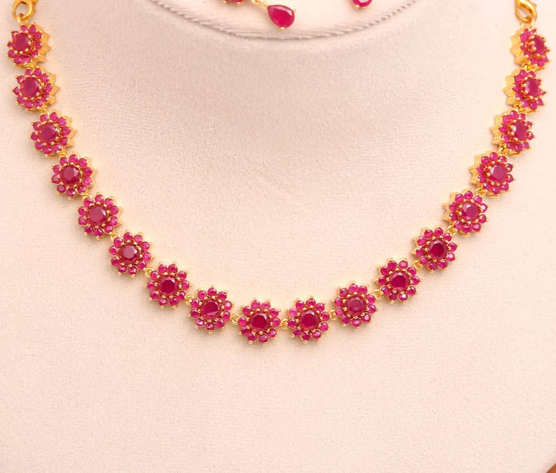 May include: A gold necklace with a floral pattern, set with bright red gemstones. The necklace is made up of flower-shaped settings, each with a central red stone and smaller red stones around it. Matching earrings are also visible.