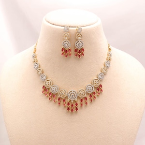 Puede incluir: Un conjunto de collar y pendientes en tono dorado con motivos circulares incrustados con piedras transparentes y acentuados por piedras rojas colgantes en forma de pera. El collar presenta un patrón repetitivo de estos elementos circulares, cada uno terminado con múltiples gotas de piedra roja a lo largo del borde inferior.