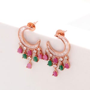 May include: Rose gold hoop earrings with a crescent design, set with small, clear crystals. The earrings have dangling teardrop-shaped gems in pink and green. The earrings have a post-back closure. The earrings are a stylish accessory.