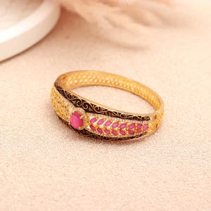 Può includere: Bracciale rigido dorato con una gemma rosa ovale centrale e gemme rosa più piccole a forma di foglia. Il bracciale presenta dettagli in smalto nero ed è adornato da piccoli cristalli scintillanti. Il bracciale ha un diametro di circa 6,35 cm.