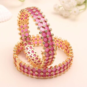 Peut inclure: Deux bracelets rigides dorés ornés d'une double rangée de pierres circulaires rose vif. Chaque pierre rose est entourée d'une petite pierre d'accent claire et scintillante sertie dans du métal doré. Les bracelets possèdent un fermoir à charnière.