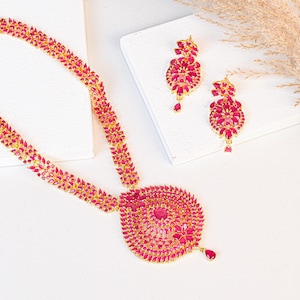 Op de afbeelding: Een gouden ketting en bijpassende oorbellen set. De ketting heeft een grote, cirkelvormige hanger en een ketting versierd met roze edelstenen. De oorbellen hebben een vergelijkbaar ontwerp, met roze edelstenen en een druppelvormige hanger.
