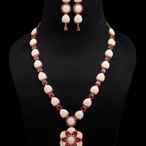 Puede incluir: Un conjunto de collar y pendientes con racimos en forma de lágrima de pequeñas cuentas blancas intercaladas con piedras rojas de corte ovalado. El collar incluye un gran colgante en forma de flor con una piedra roja central y pétalos de cuentas.