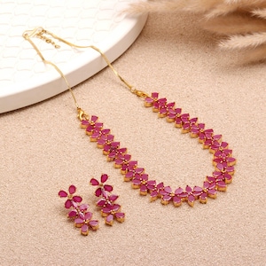 Puede incluir: Un conjunto de joyas doradas con collar y pendientes a juego. El collar y los pendientes están adornados con gemas rosas brillantes en forma de flor. El conjunto se exhibe sobre una superficie de tono neutro.