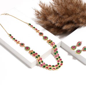 Puede incluir: Un collar dorado y un juego de pendientes a juego. El collar presenta piedras preciosas rojas y verdes alternadas en un diseño floral y ovalado. Los pendientes tienen un diseño similar. Las joyas se exhiben sobre una superficie blanca.
