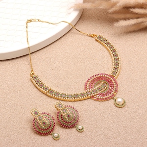 Puede incluir: Un conjunto de collar y pendientes dorados. El collar presenta un diseño en espiral con piedras preciosas rosas y verdes, y una gota de perla. Los pendientes reflejan el diseño con piedras preciosas a juego y gotas de perlas. El conjunto se muestra sobre una superficie neutra.