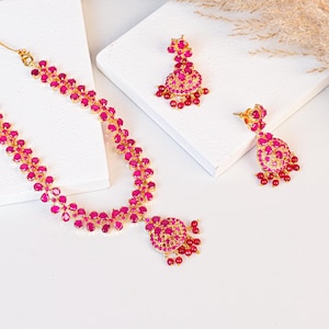 May include: A gold-coloured jewellery set comprising a necklace and earrings. The necklace and earrings are embellished with bright pink gemstones. The necklace has a floral design, and the earrings are teardrop-shaped.