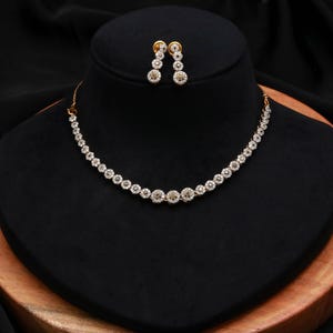 May include: A gold-toned necklace and matching pair of drop earrings, each featuring a series of circular floral-shaped links set with small, clear, sparkling stones. The necklace has a continuous row of these floral motifs, while the earrings consist of two stacked floral links each, all displayed on a dark, rounded stand.