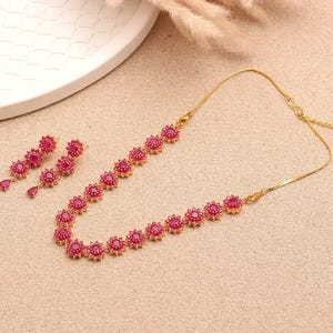 Op de afbeelding: Een goudkleurige ketting en bijpassende oorbellen set. De sieraden hebben een bloemenontwerp met rode edelstenen. De ketting en oorbellen worden gepresenteerd op een lichtbruin oppervlak, met een wit bord en gedroogde planten op de achtergrond.