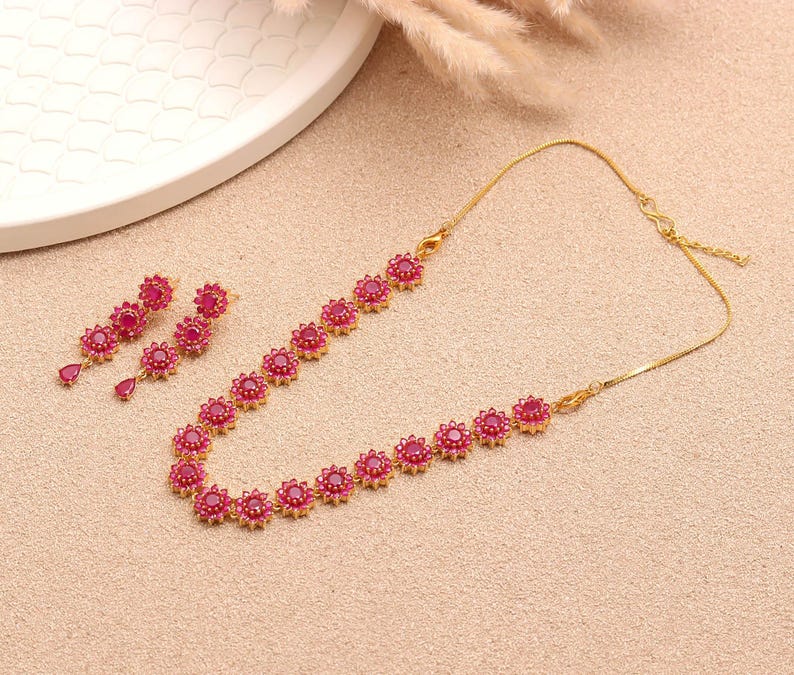 May include: A gold necklace and matching earrings set. The jewellery features a floral design with red gemstones. The necklace and earrings are displayed on a light tan surface, with a white plate and dried plants in the background.