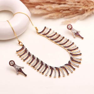 Puede incluir: Un conjunto de collar y pendientes dorados. El collar y los pendientes presentan un patrón repetido de piedras preciosas blancas y moradas alternas. Las joyas se exhiben sobre una superficie blanca con un anillo blanco decorativo y plantas secas en el fondo.