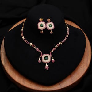 Puede incluir: Un conjunto de joyería que consta de un collar y pendientes a juego. El collar presenta un colgante cuadrado central con una piedra verde, rodeado de cristales blancos y piedras rosas, con tres gotas de piedra rosa colgantes en el centro.