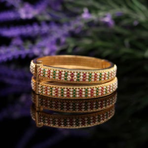 May include: Four gold bangles are stacked, reflecting on a dark surface. Each bangle is decorated with a pattern of small white, red, and green gemstones. The background features blurred purple flowers.