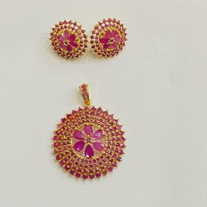 Pu&ograve; includere: Un set di gioielli color oro composto da un ciondolo circolare e due orecchini a lobo abbinati. Ogni pezzo presenta un design floreale centrale con pietre rosa, circondato da un denso alone di pietre rosa pi&ugrave; piccole su montatura dorata.