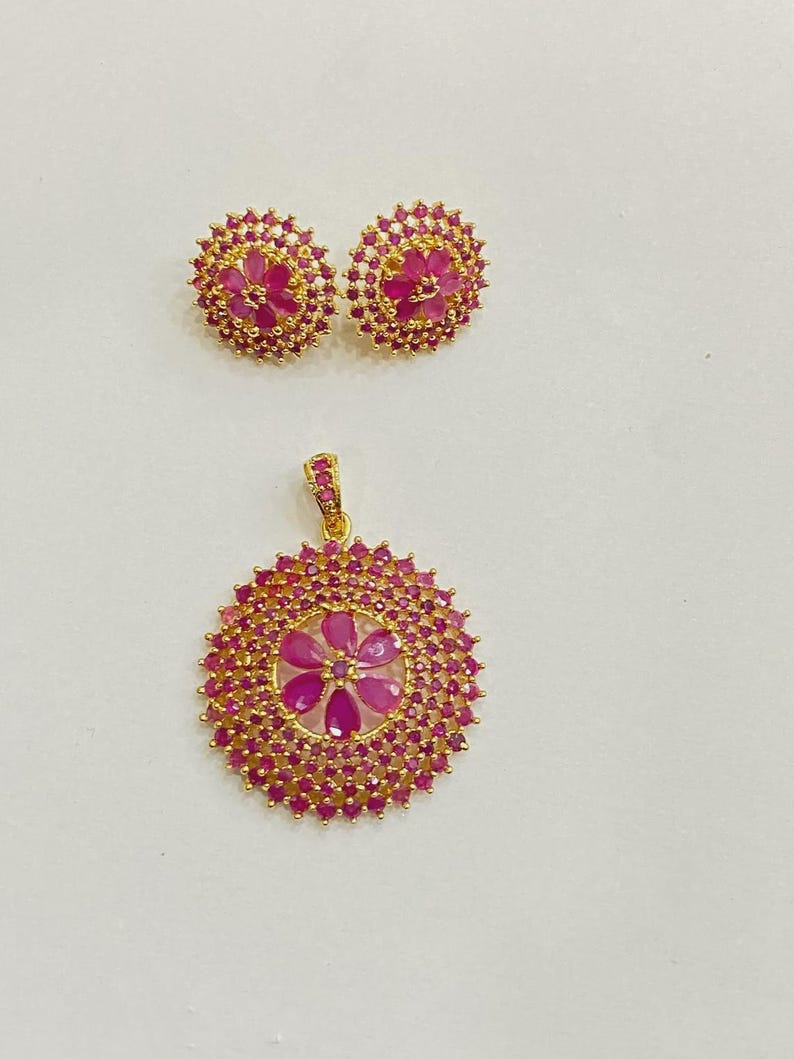 Pu&ograve; includere: Un set di gioielli color oro composto da un ciondolo circolare e due orecchini a lobo abbinati. Ogni pezzo presenta un design floreale centrale con pietre rosa, circondato da un denso alone di pietre rosa pi&ugrave; piccole su montatura dorata.