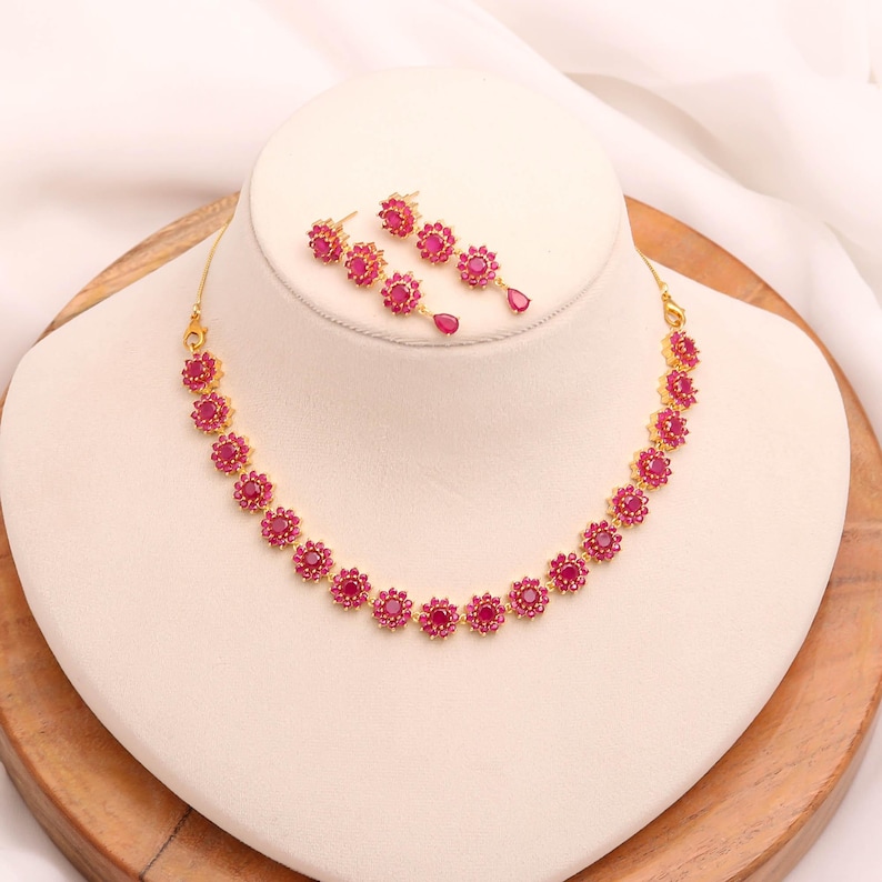 May include: A gold necklace and earring set. The necklace and earrings feature a floral design with vibrant red gemstones. The necklace is displayed on a white bust, and the earrings are arranged to the side. The jewellery is set against a neutral background.