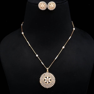 Può includere: Un set composto da collana e orecchini a lobo color oro, con un ciondolo circolare e orecchini caratterizzati da un motivo floreale centrale circondato da pietre bianche. La catenina è impreziosita da piccole perline bianche e distanziatori cilindrici.