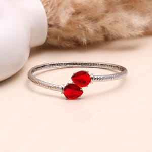 May include: A silver bangle bracelet featuring two bright red, teardrop-shaped gemstones. The bracelet has an engraved floral pattern along the band. The gemstones are set in a bezel setting.