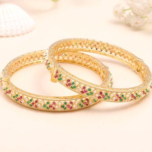 May include: Two gold-colored bangles with intricate designs. Each bangle is adorned with small, colorful gemstones in red, green, and white. The bangles are set against a soft, neutral background, highlighting their ornate details.