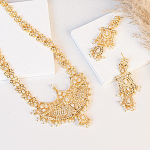 May include: A gold-toned jewelry set featuring a necklace and earrings. The necklace has a floral design with small pearls, leading to a fan-shaped pendant. The earrings mirror the design with pearl accents. The set is displayed on a white surface.