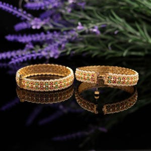 Könnte beinhalten: Zwei goldfarbene Armreifen mit aufwendigen Details. Jeder Armreif ist mit kleinen, farbigen Edelsteinen in Rot, Grün und Weiß verziert, die auf einem goldenen Hintergrund gesetzt sind. Die Armreifen werden auf einer reflektierenden Oberfläche mit verschwommenem floralen Hintergrund gezeigt.