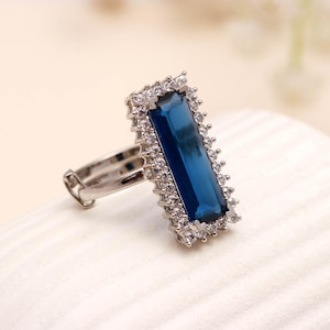May include: A silver-toned ring featuring a rectangular, deep blue gemstone. The gemstone is surrounded by a halo of smaller, clear stones. The ring's band is adjustable, and the design is elegant and eye-catching.