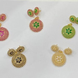 Pu&ograve; includere: Cinque set di pendenti circolari e orecchini a lobo abbinati in vari colori. Ogni set presenta un motivo floreale centrale circondato da cerchi concentrici di piccole pietre. I colori includono rosa, verde, nero e bianco con metallo dorato.