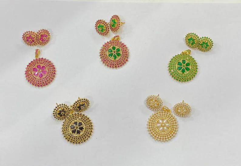 Pu&ograve; includere: Cinque set di pendenti circolari e orecchini a lobo abbinati in vari colori. Ogni set presenta un motivo floreale centrale circondato da cerchi concentrici di piccole pietre. I colori includono rosa, verde, nero e bianco con metallo dorato.