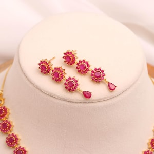 May include: Gold-coloured jewellery set featuring a necklace and earrings. The earrings are a drop style with a floral design and a teardrop-shaped ruby. The necklace has a matching floral design with ruby-coloured stones.