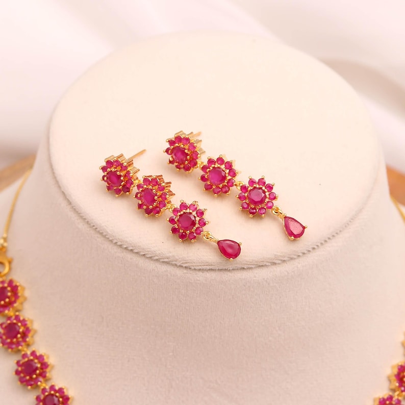 May include: Gold-coloured jewellery set featuring a necklace and earrings. The earrings are a drop style with a floral design and a teardrop-shaped ruby. The necklace has a matching floral design with ruby-coloured stones.