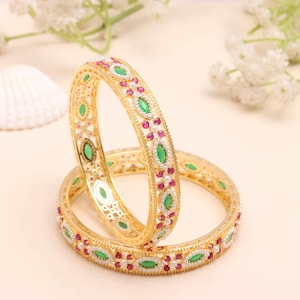 Puede incluir: Dos brazaletes dorados con diseños intrincados. Cada brazalete presenta gemas verdes y rojas alternas, con pequeños detalles blancos. Los brazaletes se colocan sobre un fondo suave y neutro, resaltando sus detalles ornamentales.