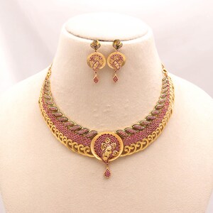 Puede incluir: Un collar dorado y un par de pendientes a juego con intrincados trabajos de filigrana metálica e incrustaciones de pequeñas piedras rosas. El collar tiene un diseño curvo en forma de media luna con un colgante circular central y una gota.
