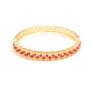 K&ouml;nnte beinhalten: Ein goldfarbenes Armreif-Armband mit einer Reihe kleiner, runder, roter Edelsteine. Das Armband hat ein filigranes Design mit einer Reihe verbundener Kreise, die die Edelsteine halten. Das Armband hat ein geometrisches Muster.