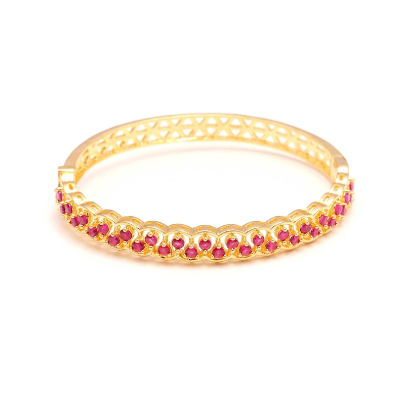 K&ouml;nnte beinhalten: Ein goldfarbenes Armreif-Armband mit einer Reihe kleiner, runder, roter Edelsteine. Das Armband hat ein filigranes Design mit einer Reihe verbundener Kreise, die die Edelsteine halten. Das Armband hat ein geometrisches Muster.