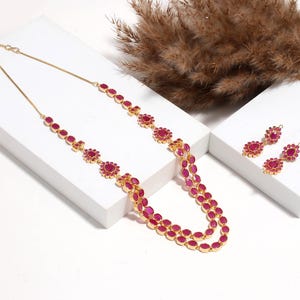 Puede incluir: Un conjunto de collar y pendientes dorados. El collar presenta un diseño de doble hilera con piedras preciosas de color rubí de forma ovalada y floral. Los pendientes tienen un diseño floral similar con piedras de color rubí. La joyería se exhibe sobre una superficie blanca.