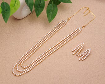 Könnte beinhalten: Ein goldfarbenes Schmuckset mit einer langen Halskette und passenden Ohrringen. Die Halskette und die Ohrringe sind mit zahlreichen kleinen, funkelnden weißen Steinen verziert. Das Set wird auf einer neutral getönten Oberfläche präsentiert, mit einer weißen Vase und grünen Blättern im Hintergrund.