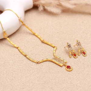 May include: A gold-coloured jewellery set comprising a necklace and matching earrings. The necklace features a pendant with a red gemstone. Both the earrings and necklace are embellished with small, clear gemstones. The set is presented on a neutral-toned surface.