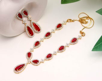 Puede incluir: Un conjunto de joyas de tono dorado con un collar y pendientes a juego. El collar y los pendientes tienen piedras preciosas rojas en forma de lágrima, cada una rodeada por un halo de pequeñas piedras transparentes. El conjunto se exhibe sobre una superficie blanca.