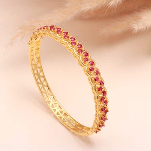 Puede incluir: Una pulsera dorada con una fila de pequeñas piedras preciosas rojas redondas. La pulsera tiene un diseño geométrico en los laterales y una serie de piedras preciosas rojas en la parte superior. La joya se exhibe sobre un fondo rosa claro.
