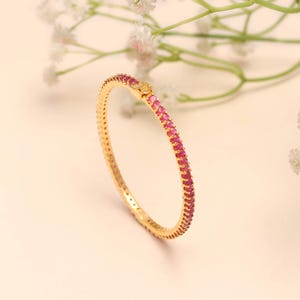 Può includere: Un braccialetto dorato con una fila di piccole pietre preziose rosa brillante lungo la metà superiore. Il braccialetto è circolare e ha un design semplice ed elegante. Lo sfondo è di un colore neutro e morbido con fiori bianchi.