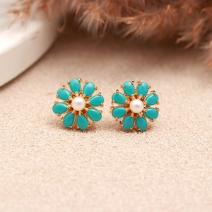 May include: A pair of gold-tone stud earrings shaped like flowers. Each earring features a central white pearl surrounded by ten teardrop-shaped turquoise-coloured stones arranged in a circular pattern, set in a polished gold-coloured metal frame.