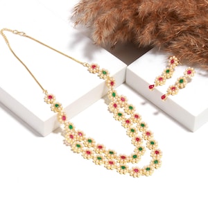 Pode incluir: Um conjunto de colar e brincos dourados. O colar apresenta uma dupla fileira de pequenas pérolas brancas e detalhes em forma de flor com pedras preciosas vermelhas e verdes. Os brincos têm um design semelhante com uma gema vermelha em forma de gota.