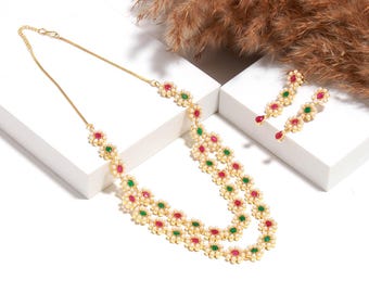 May include: A gold-toned necklace and matching earrings set. The necklace features a double strand of small, white pearls and flower-shaped accents with red and green gemstones. The earrings have a similar design with a teardrop-shaped red gemstone.