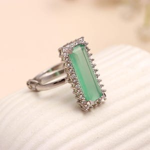Peut inclure: Une bague en argent ornée d'une pierre verte pâle rectangulaire, entourée de petites pierres claires. Le corps de la bague présente un design simple et élégant, idéal pour toutes les occasions.