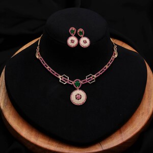 Puede incluir: Un conjunto de joyería compuesto por un collar y pendientes a juego. El collar presenta una cadena con piedras rosas, eslabones geométricos dorados, una piedra verde central en forma de lágrima y un colgante circular con perlas blancas.